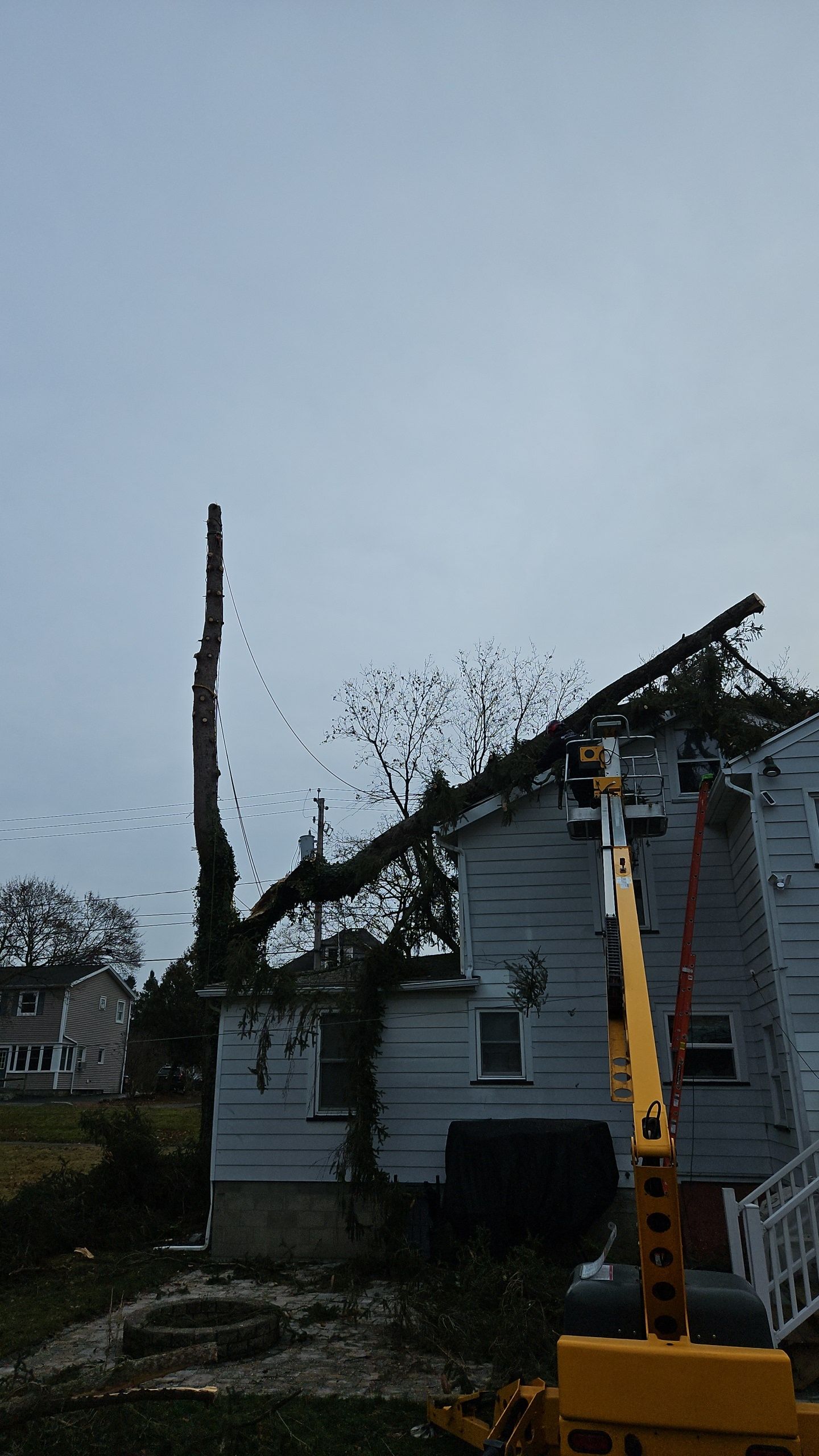 A yellow boom lift clears a fallen tree branch from the roof of a grey two-story house.