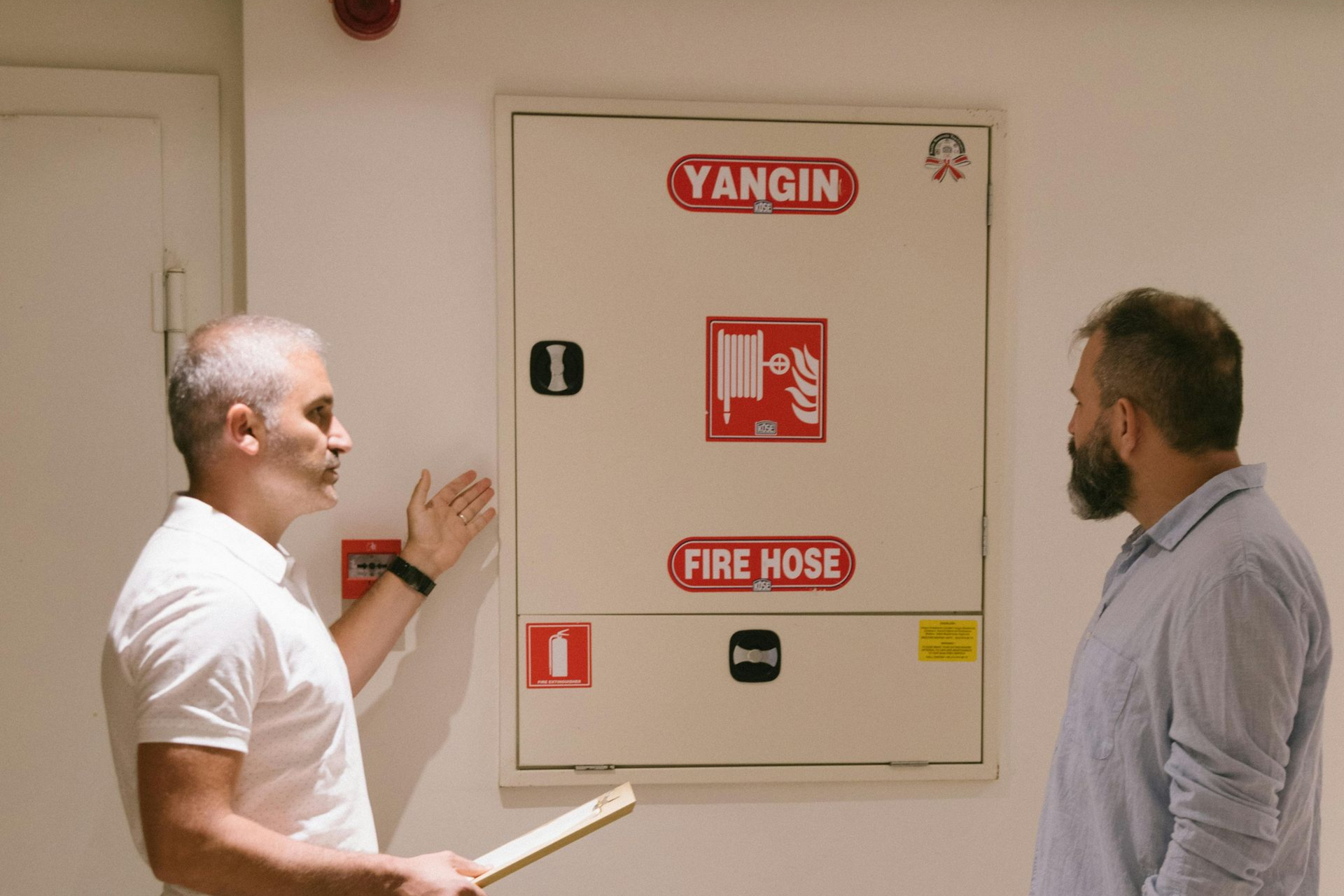 Two men inspect a wall-mounted fire hose cabinet labeled “YANGIN” and “FIRE HOSE.”