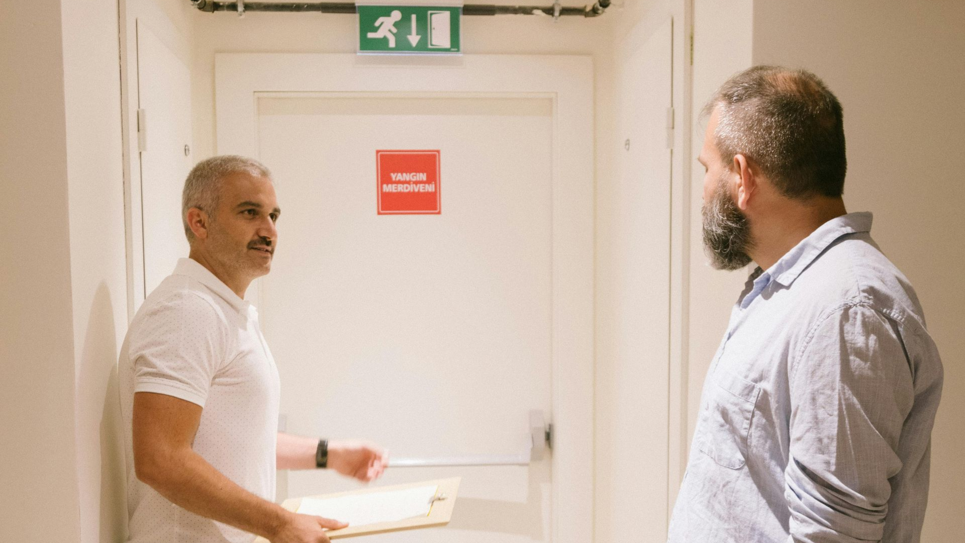 Two men in a bright hallway, one holding a tray near a white door with an exit sign above it.