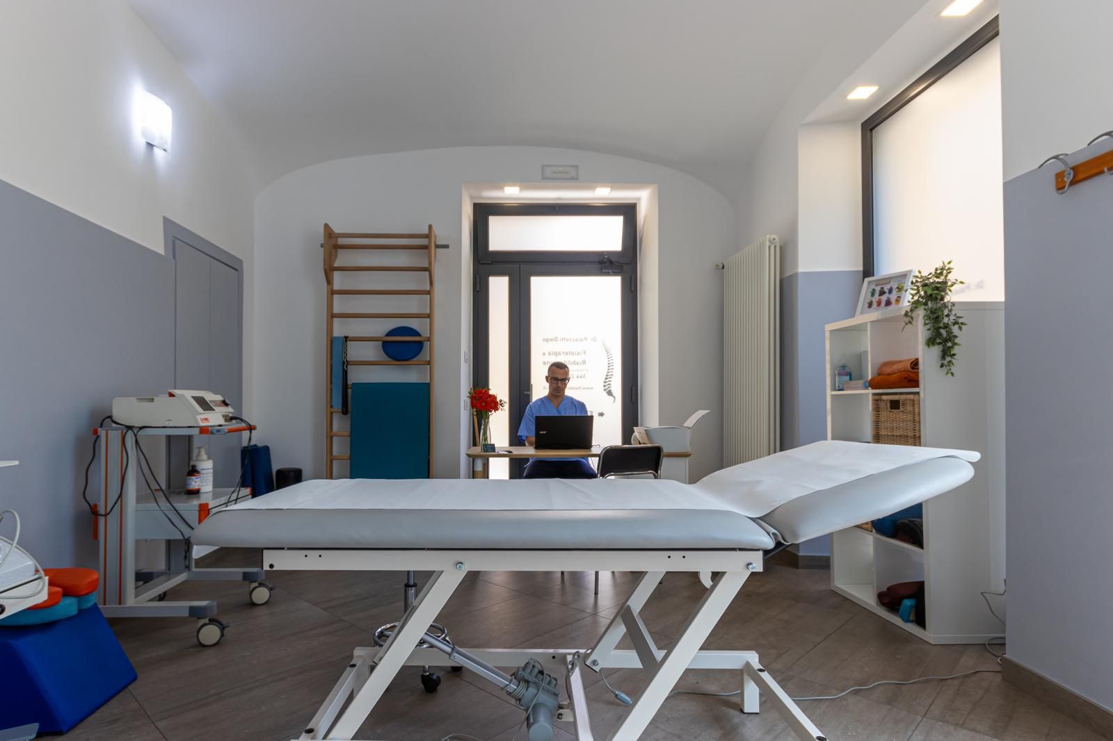 Stanza vuota per fisioterapia con due lettini per trattamenti e attrezzi per esercizi vicino a una porta.