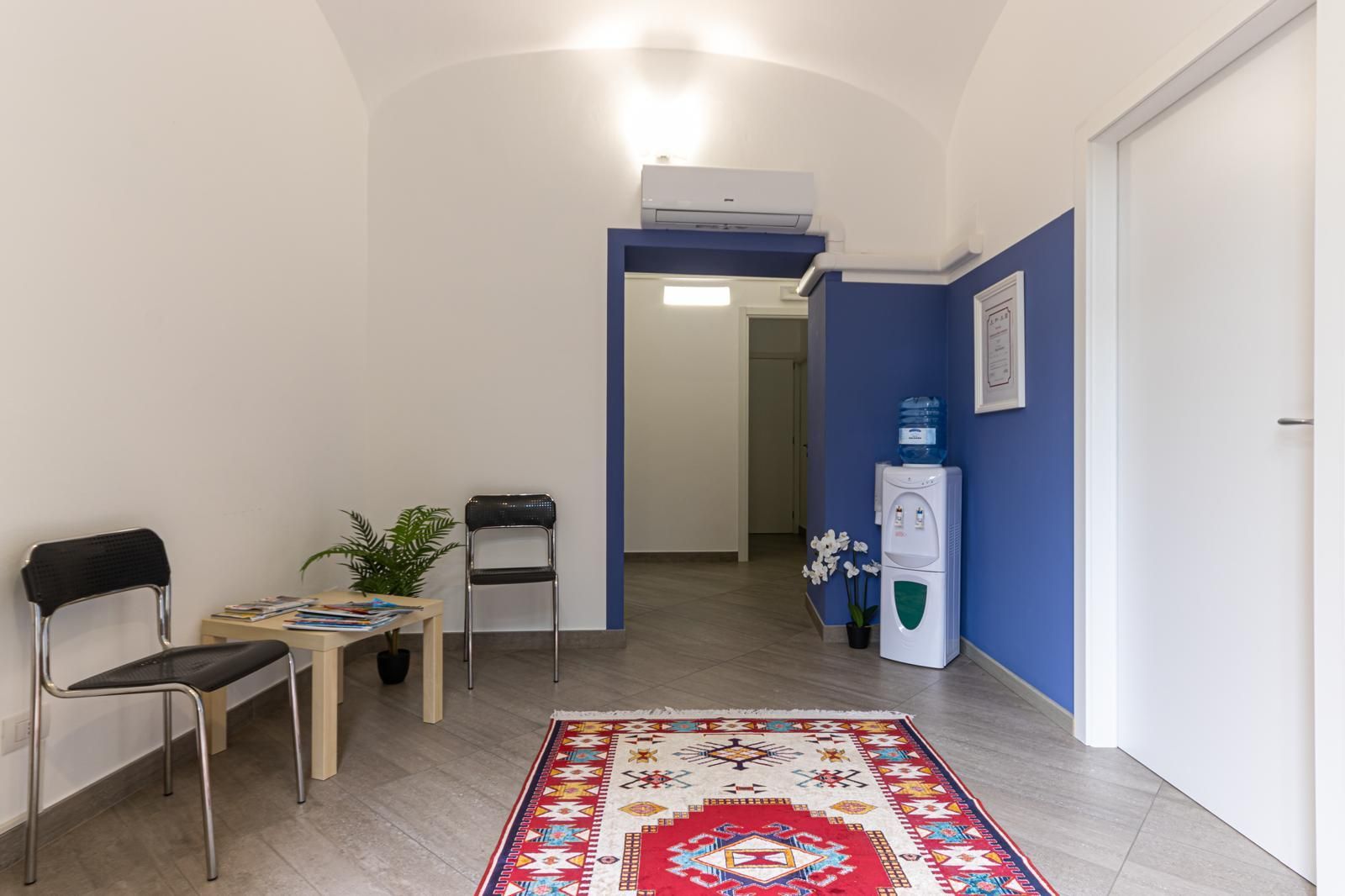 Piccola sala d'attesa con sedie, tavolo, pianta, distributore d'acqua e parete blu a contrasto, vista in fondo a un corridoio.