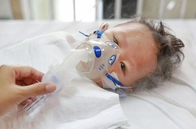 Baby lying in bed wearing an oxygen mask, with a caregiver’s hand adjusting it