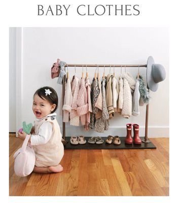 Baby clothes on a rack beside a smiling toddler in a white outfit on a wooden floor.