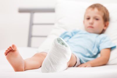 Child with a bandaged foot lying on a bed, looking upset in a bright room