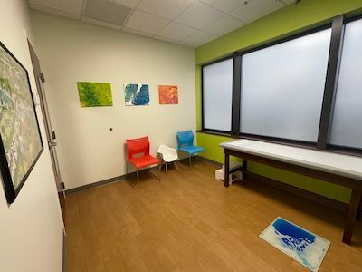 Small waiting room with two chairs, desk by frosted window, colorful wall art, and floor mat.