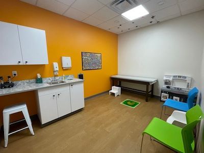 Clinic exam room with sink, table, scale, and colorful chairs on orange and white walls