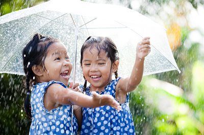 Two children smiling under a clear umbrella in the rain, wearing blue dresses.