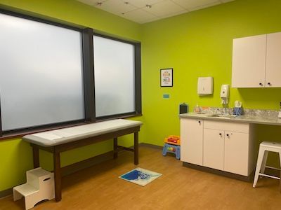 Bright pediatric exam room with green walls, exam table, sink, cabinets, and child stool.
