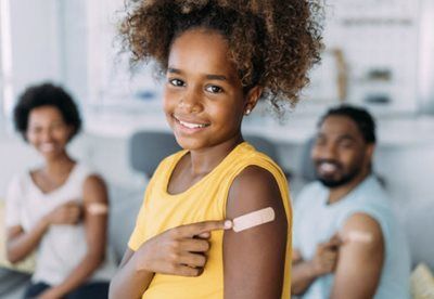 Smiling child showing a bandaged upper arm in a bright room, with two blurred people in the background
