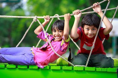Two children smiling on a green rope bridge, holding overhead ropes outdoors.