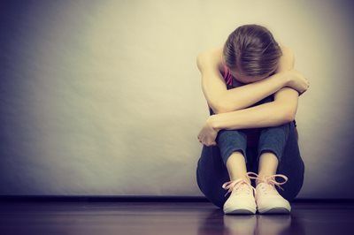 Person sitting on the floor with arms wrapped around knees, head down in a dim room