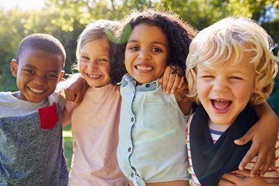 Four smiling children outdoors, leaning together in a sunny park.
