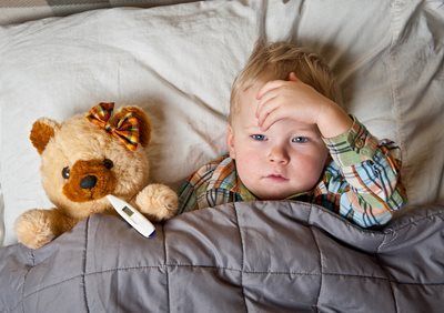Child lying in bed with hand on forehead beside a teddy bear under a gray blanket