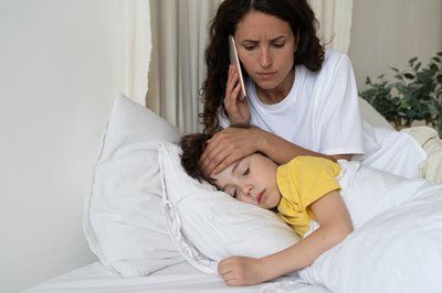 Woman comforting sick child in bed, holding a thermometer to her forehead.