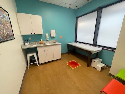 Small exam room with sink, cabinets, stool, bench, and a window; orange mat on the floor.