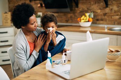 Woman helping a child blow their nose beside a laptop in a home kitchen.