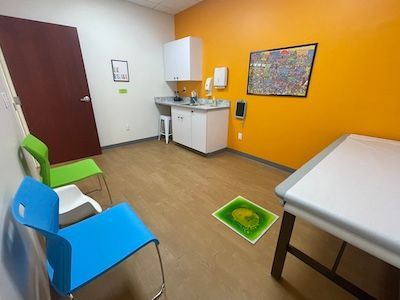 Exam room with chairs, sink cabinet, and exam table; bright orange and white walls.