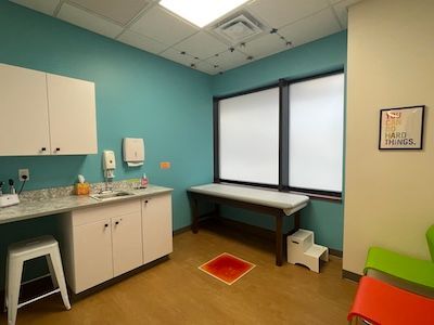 Small medical exam room with sink, cabinets, exam table, window, and colorful chairs.