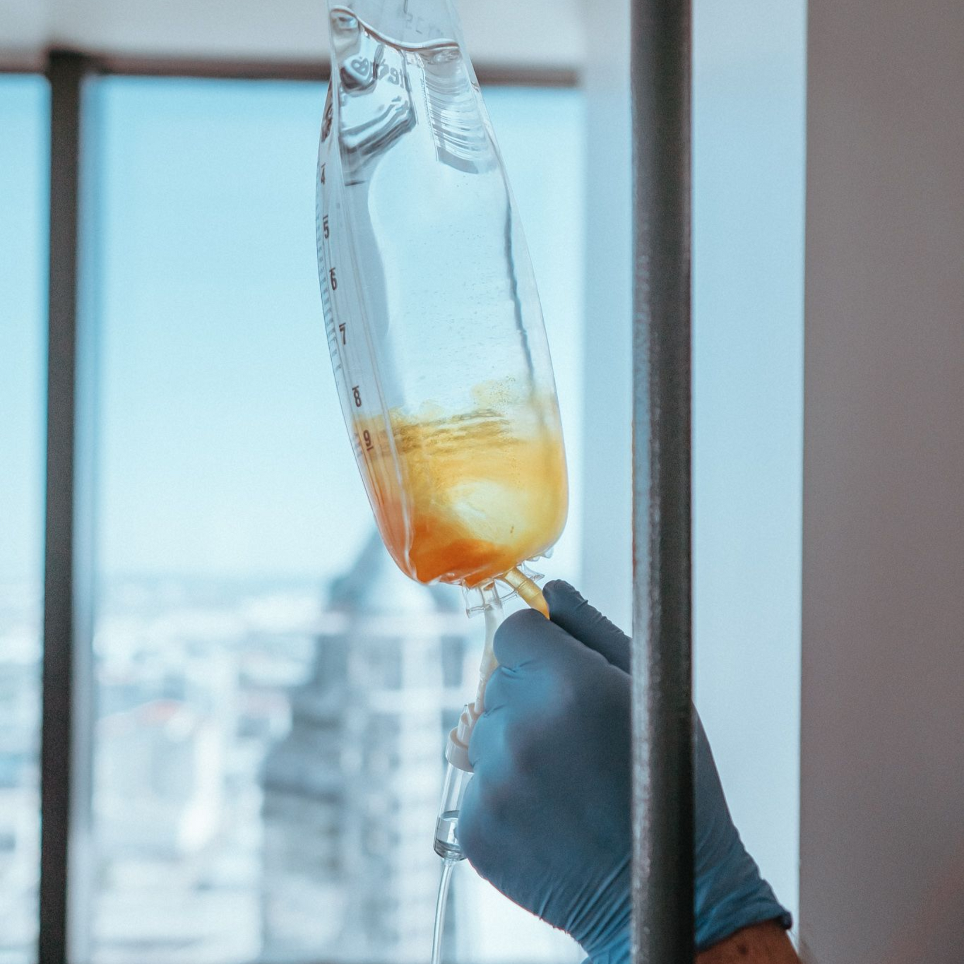 Hand with a blue glove adjusting an IV drip bag containing a yellow liquid against a window.