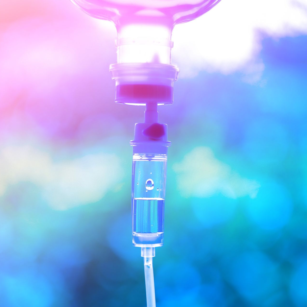 IV drip bag with fluid, connected to tubing. Pink and blue bokeh background.