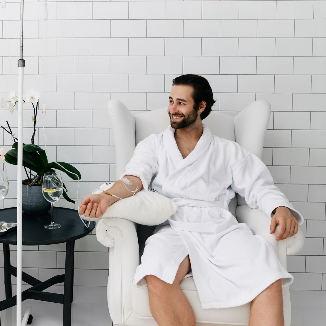 Man in white robe smiles while receiving IV drip in white-tiled room.