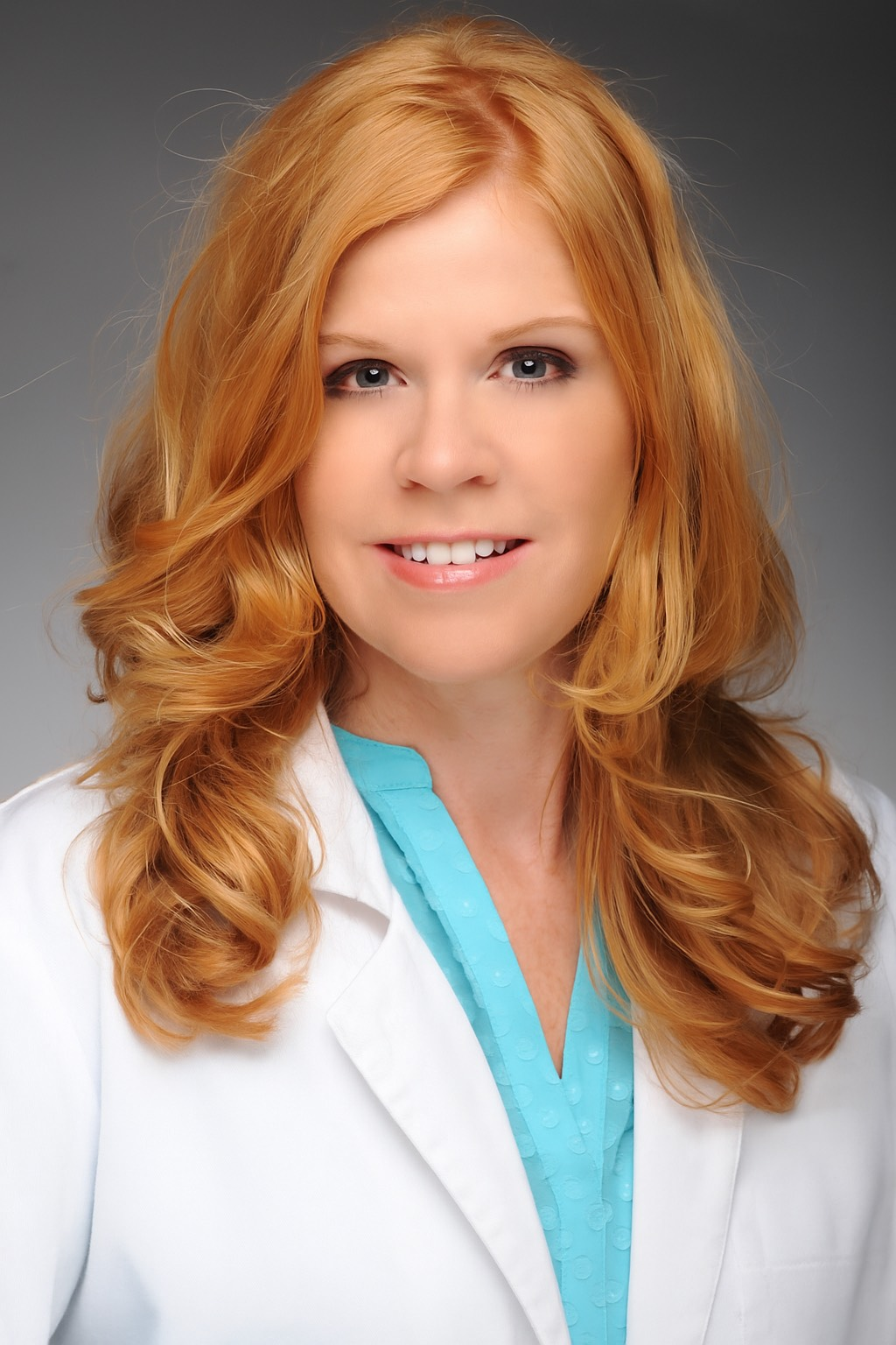 Woman with red hair smiling, wearing a white coat over a blue top, against a gray gradient background.