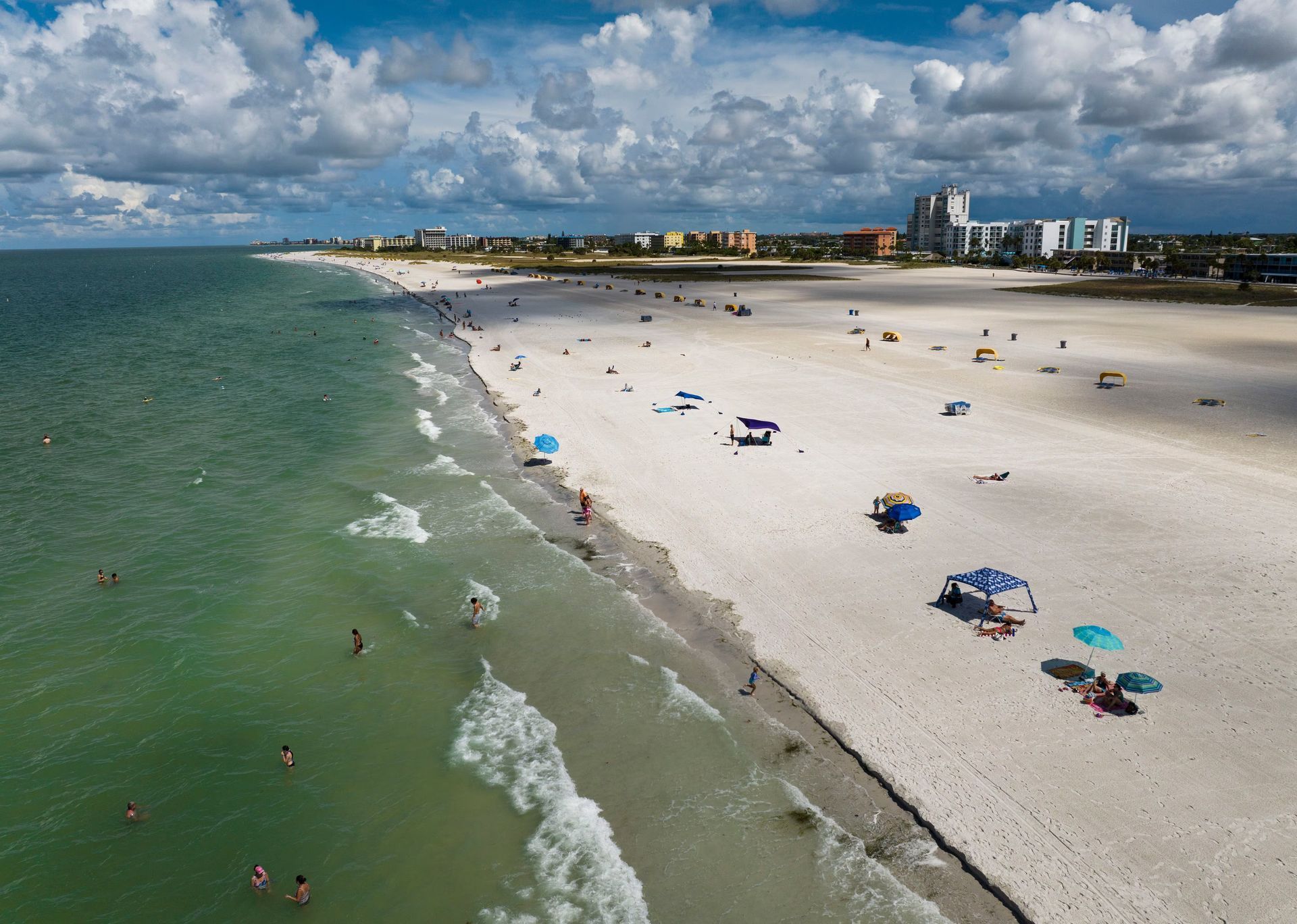 Beach with white sand, turquoise water, and people swimming and sunbathing under umbrellas on a sunny day.