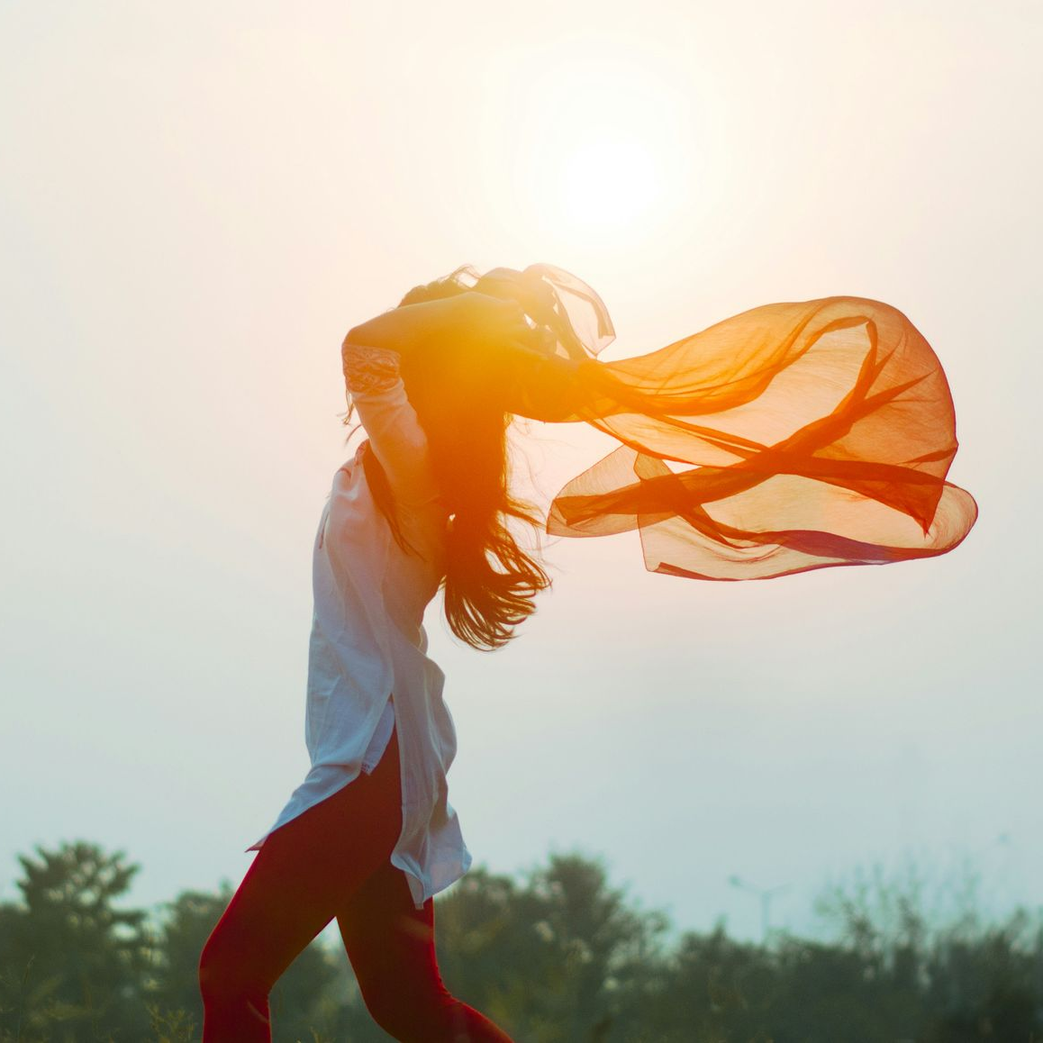 Woman with long hair and flowing orange scarf, reaching upward, bright sunlight.
