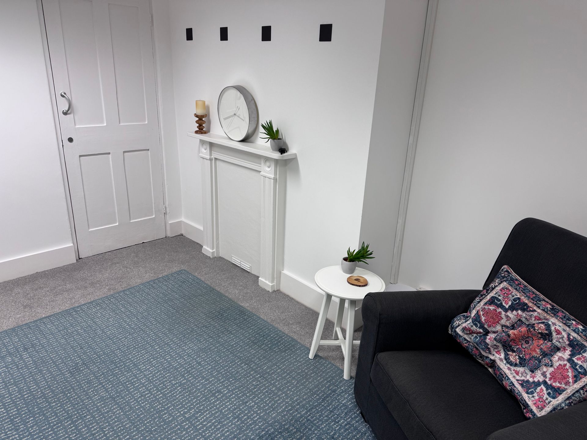 Small living room with gray carpet, white mantel, black sofa, and a door near the wall.