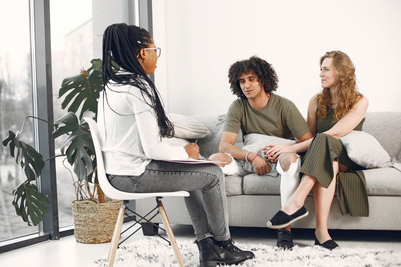 Three people talking on a sofa in a bright living room, with a potted plant and woven basket nearby.