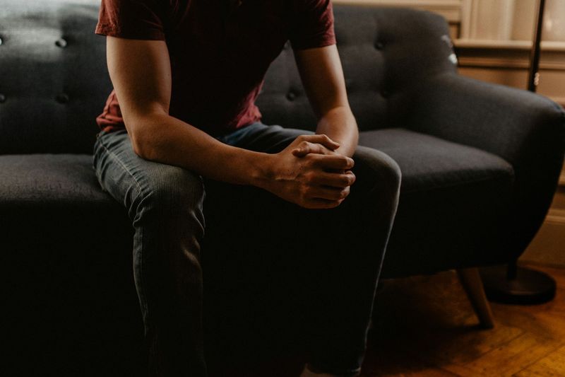 Person seated on a dark couch, hands clasped, in a dimly lit living room.