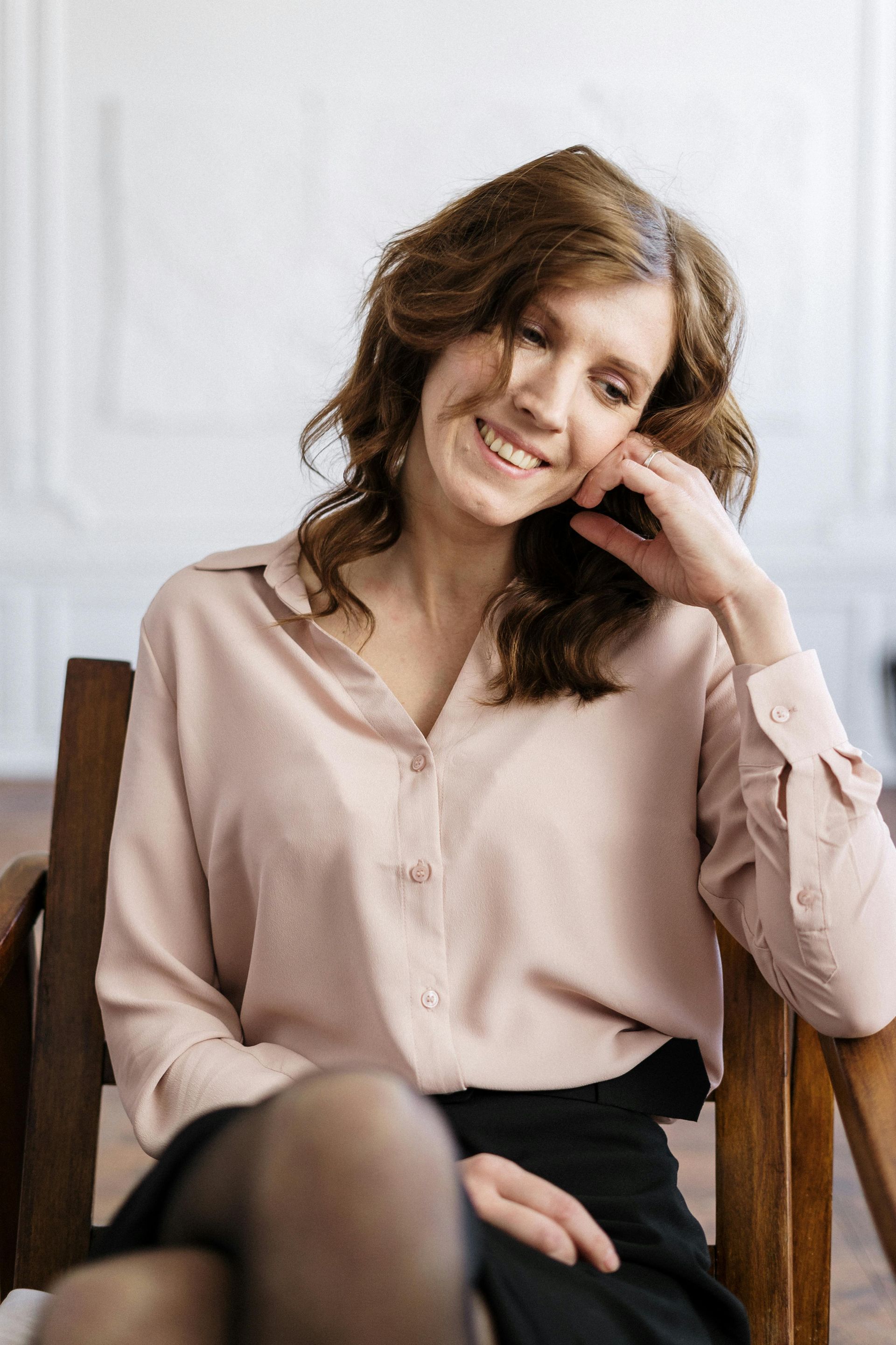 Smiling woman in a light pink blouse seated by a window, resting her head on her hand.
