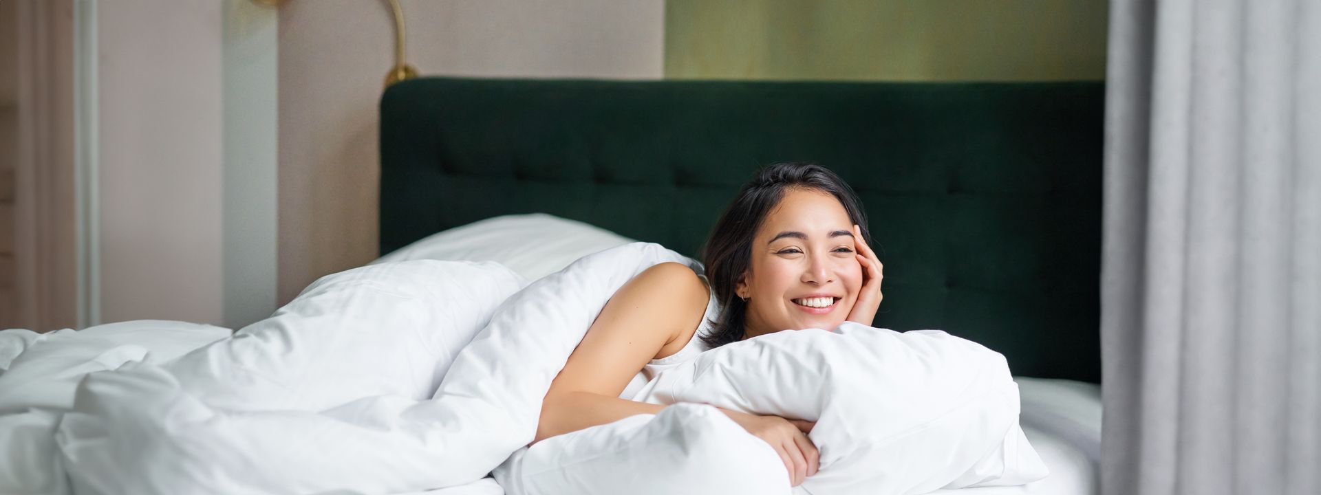 一位女子微笑著躺在鋪著白色床單的床上，臥室燈光柔和。
