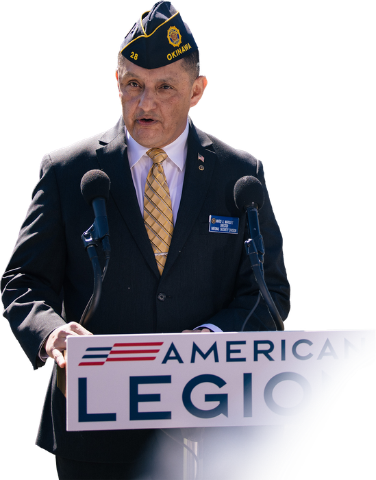 A person in a dark suit and American Legion cap speaks at a podium with an American Legion sign.