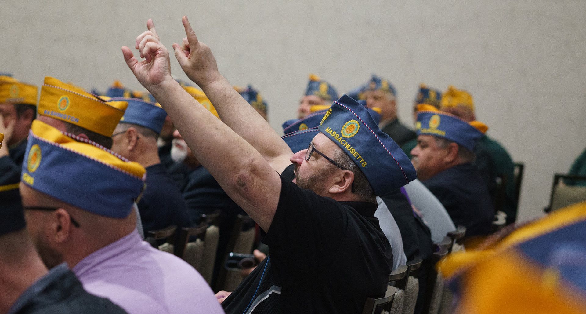 A man in a blue military-style cap raises both arms with index fingers pointed up during an indoor meeting.
