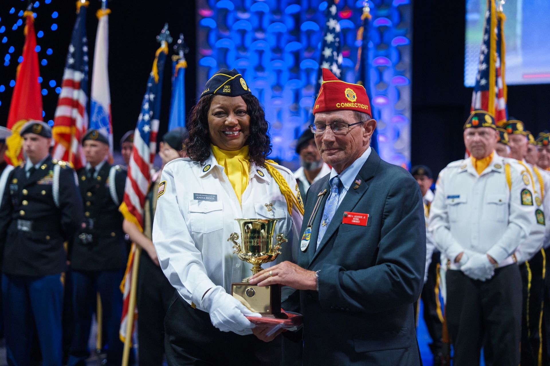 A smiling woman and a man in a veteran's cap hold a trophy together on a stage with flags behind them.