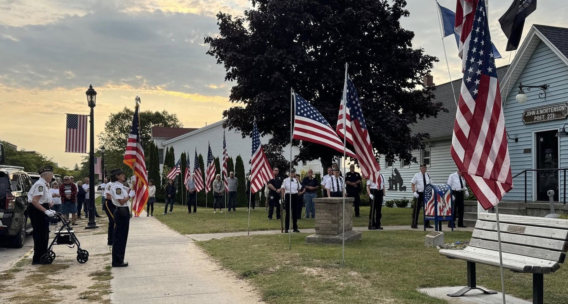 A group of people standing on a lawn at sunset, holding many American flags during a commemorative outdoor ceremony.
