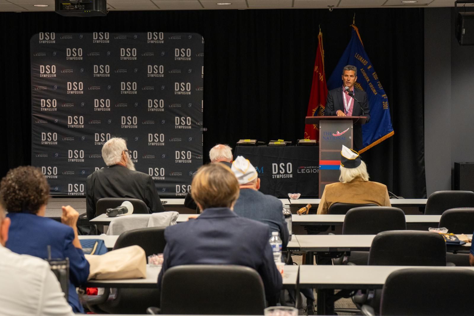 A speaker stands at a podium addressing an audience in a conference room with a branded backdrop and two flags.