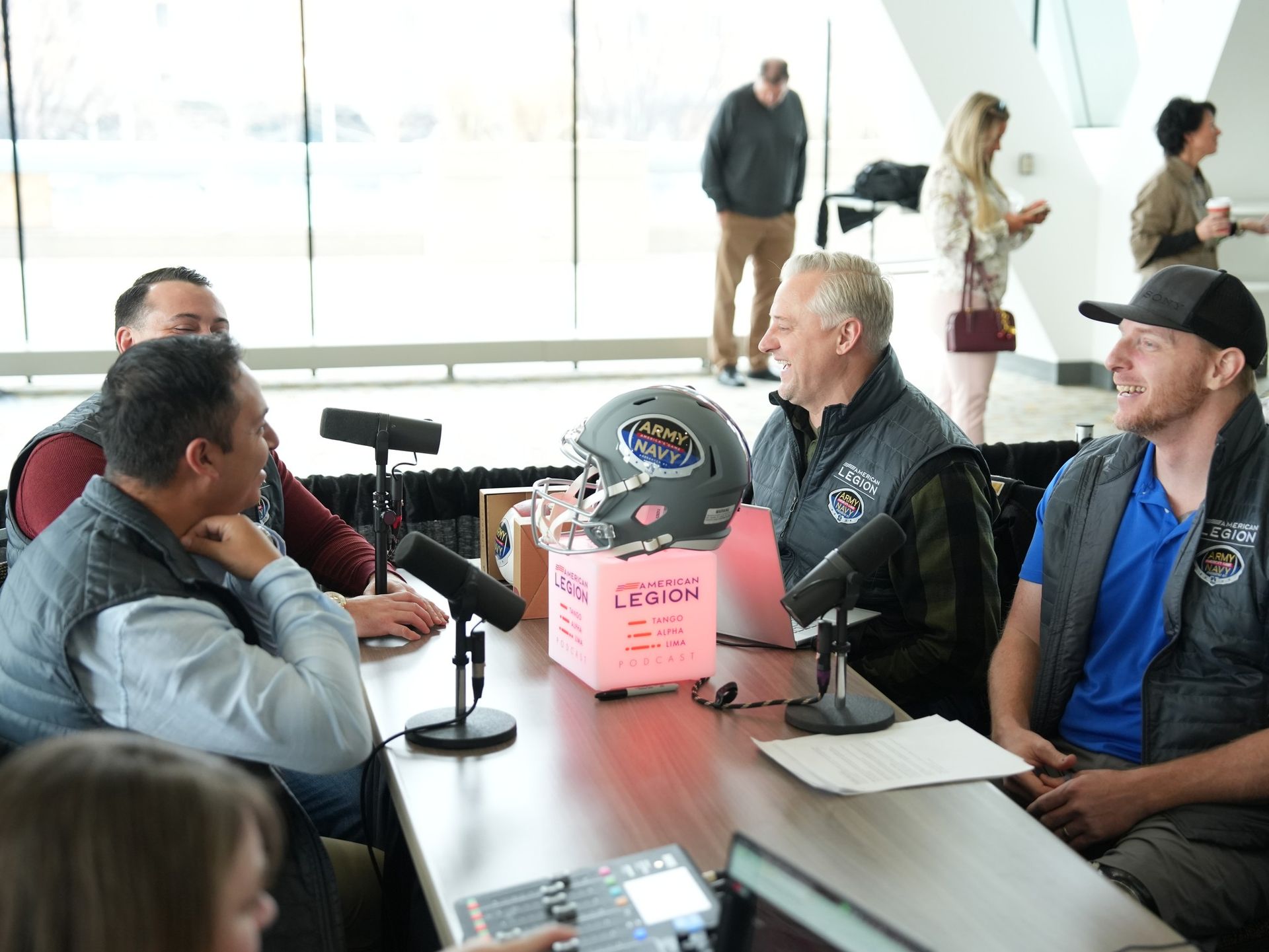 Four people sit at a table with microphones and a sports helmet for a podcast recording in a brightly lit, open room.