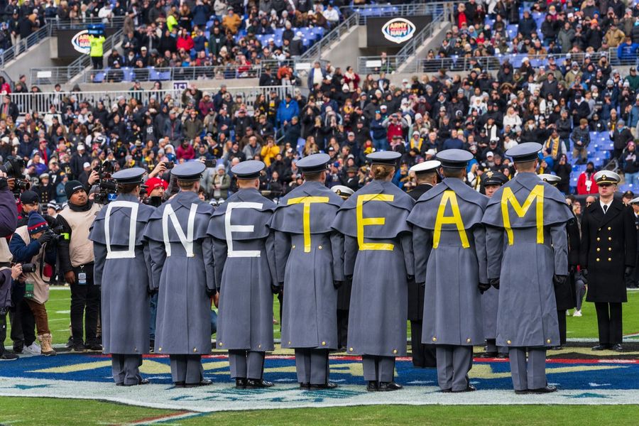 Uniformed cadets stand in a row on a football field, their coats displaying the yellow letters