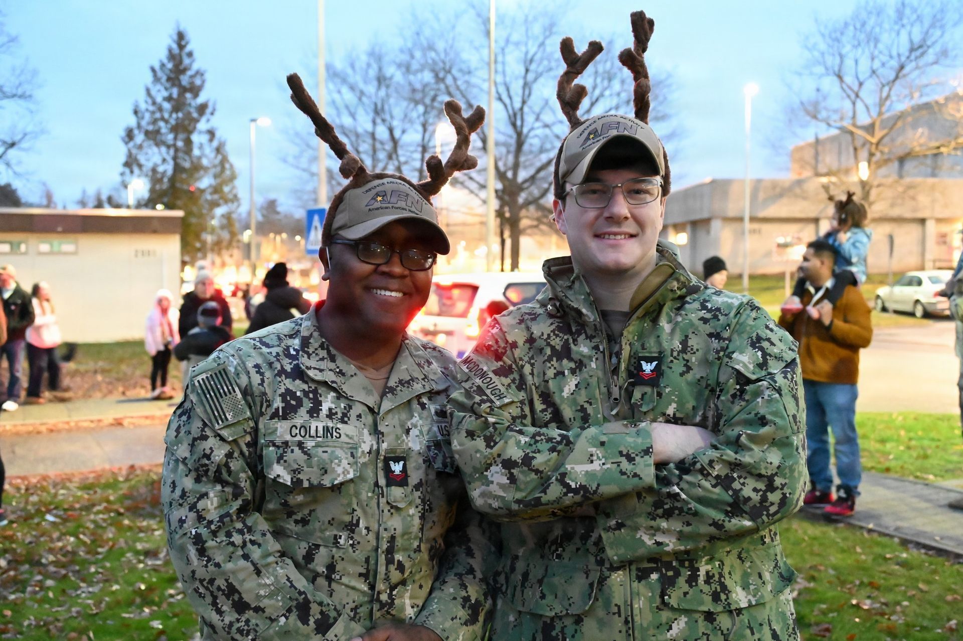 Two people in camouflage uniforms wearing reindeer antler headbands, smiling outdoors in a park setting.