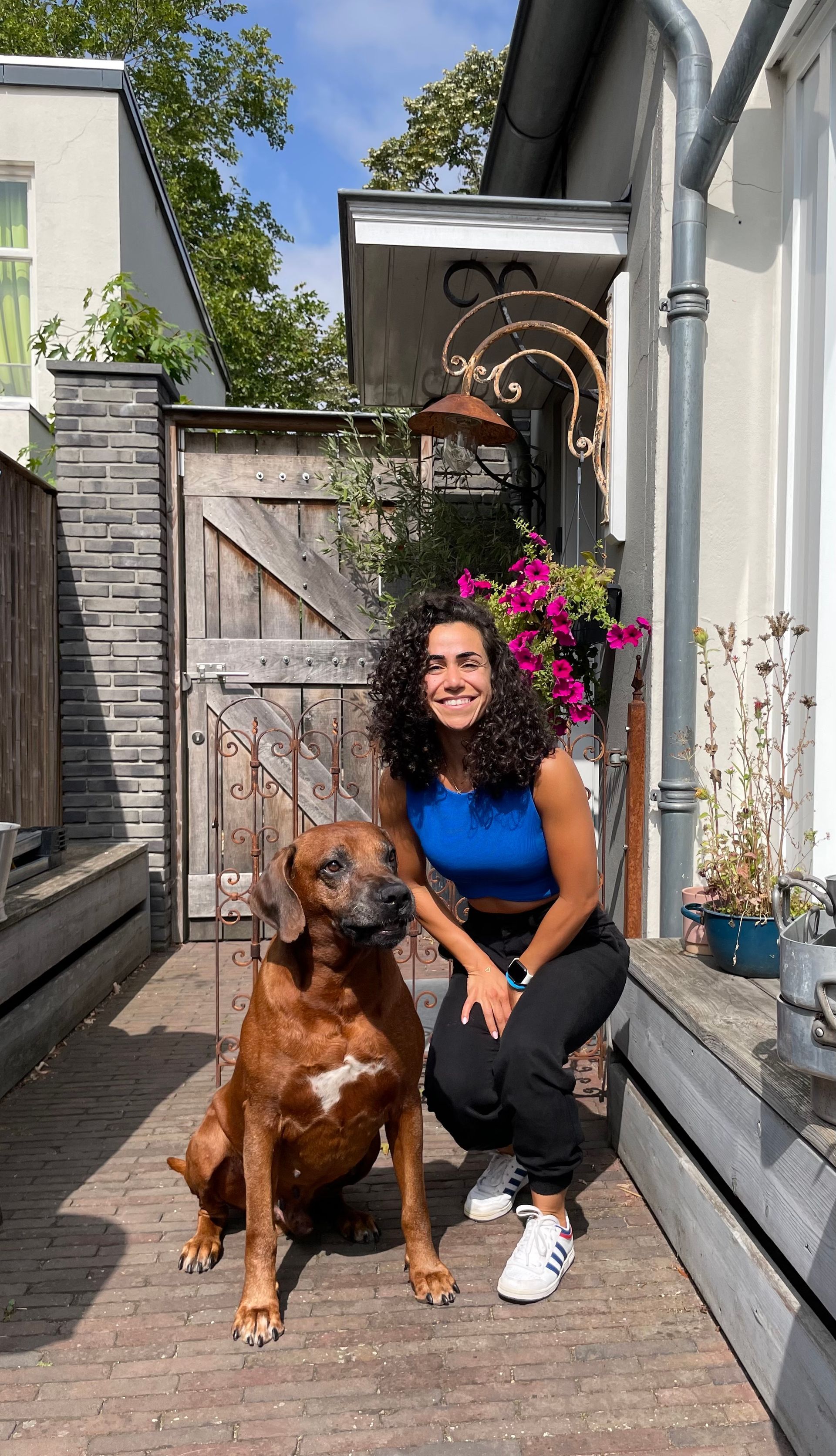 Vrouw zit met een bruine hond op een houten terras naast een rustieke schuur en bloemen.