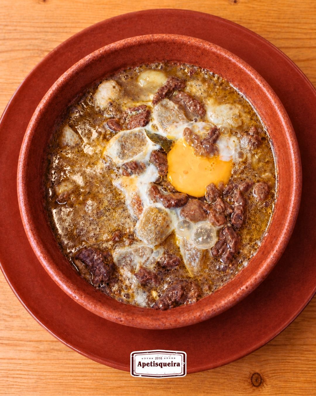 Tigela de barro vermelho com ensopado assado e queijo derretido sobre uma mesa de madeira