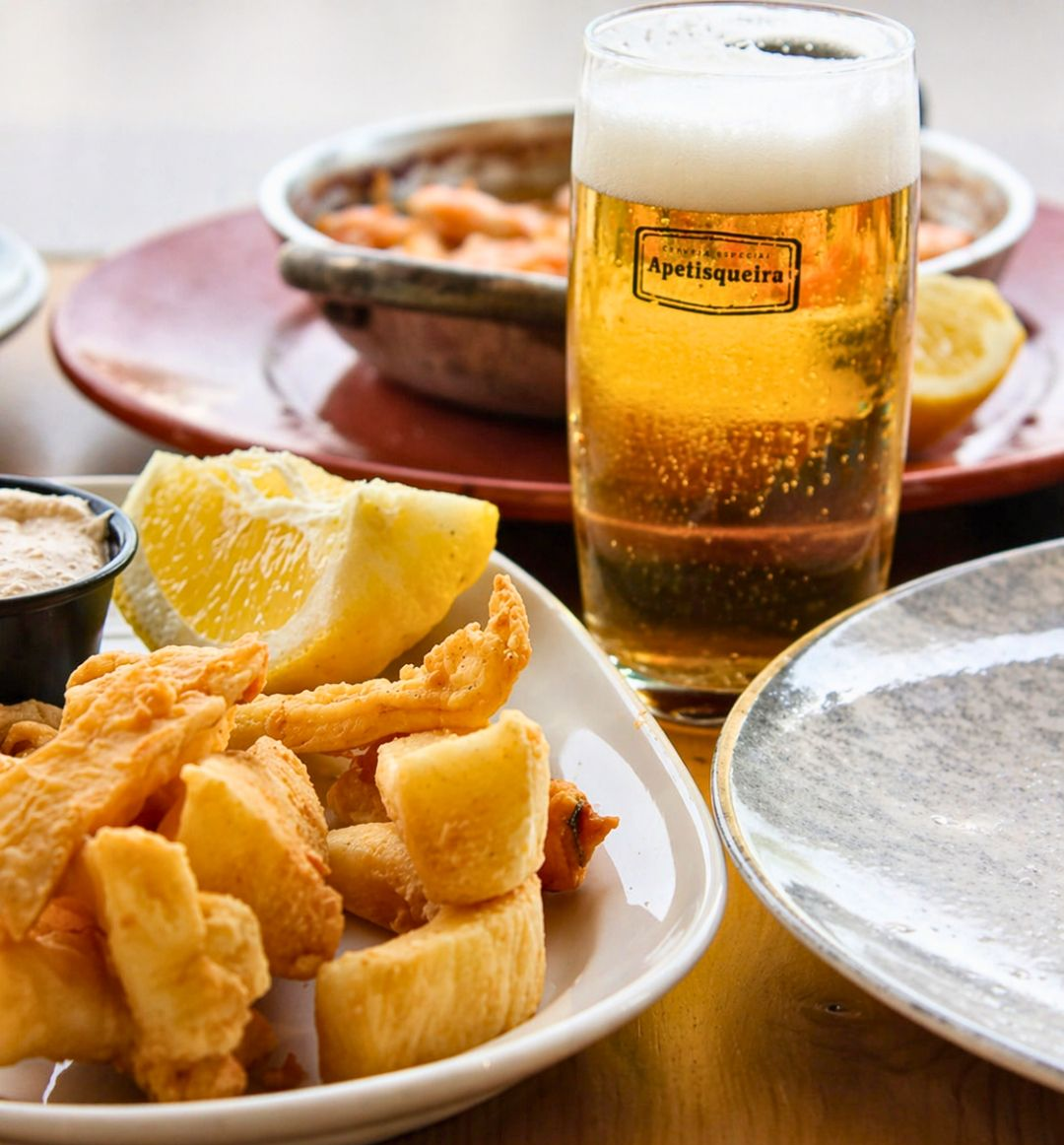 Copo de cerveja com limão e petiscos fritos em pratos sobre uma mesa.