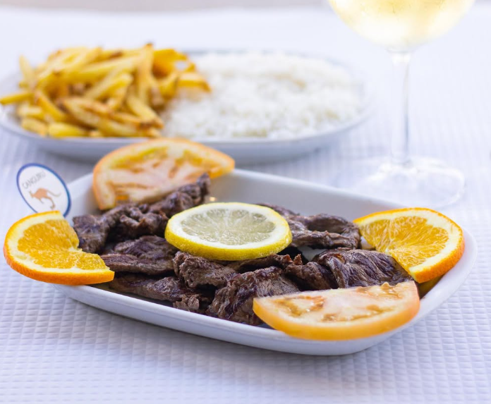 Bife grelhado com fatias de laranja e limão em um prato branco, com batatas fritas e arroz ao fundo.