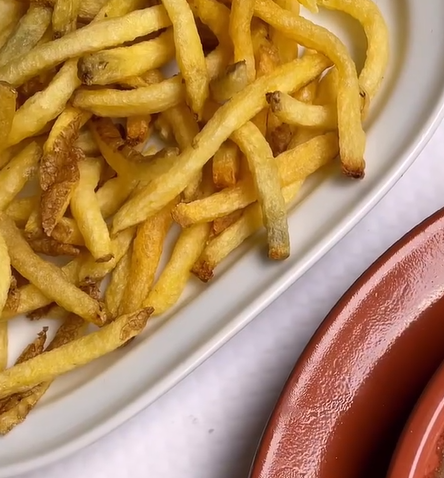Prato de batatas fritas douradas ao lado de um molho de terracota sobre uma mesa branca.