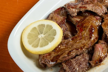 Carne grelhada em um prato branco com uma fatia de limão por cima, sobre uma toalha de mesa laranja.