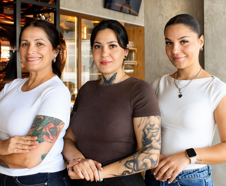 Três mulheres em pé juntas dentro de casa, sorrindo, com tatuagens e roupas casuais.