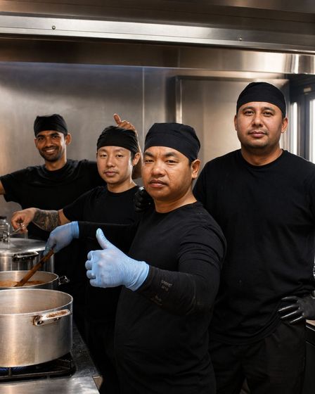 Quatro chefs de uniforme e chapéu pretos estão em uma cozinha; um deles usa luvas azuis e faz um sinal de positivo com o polegar.