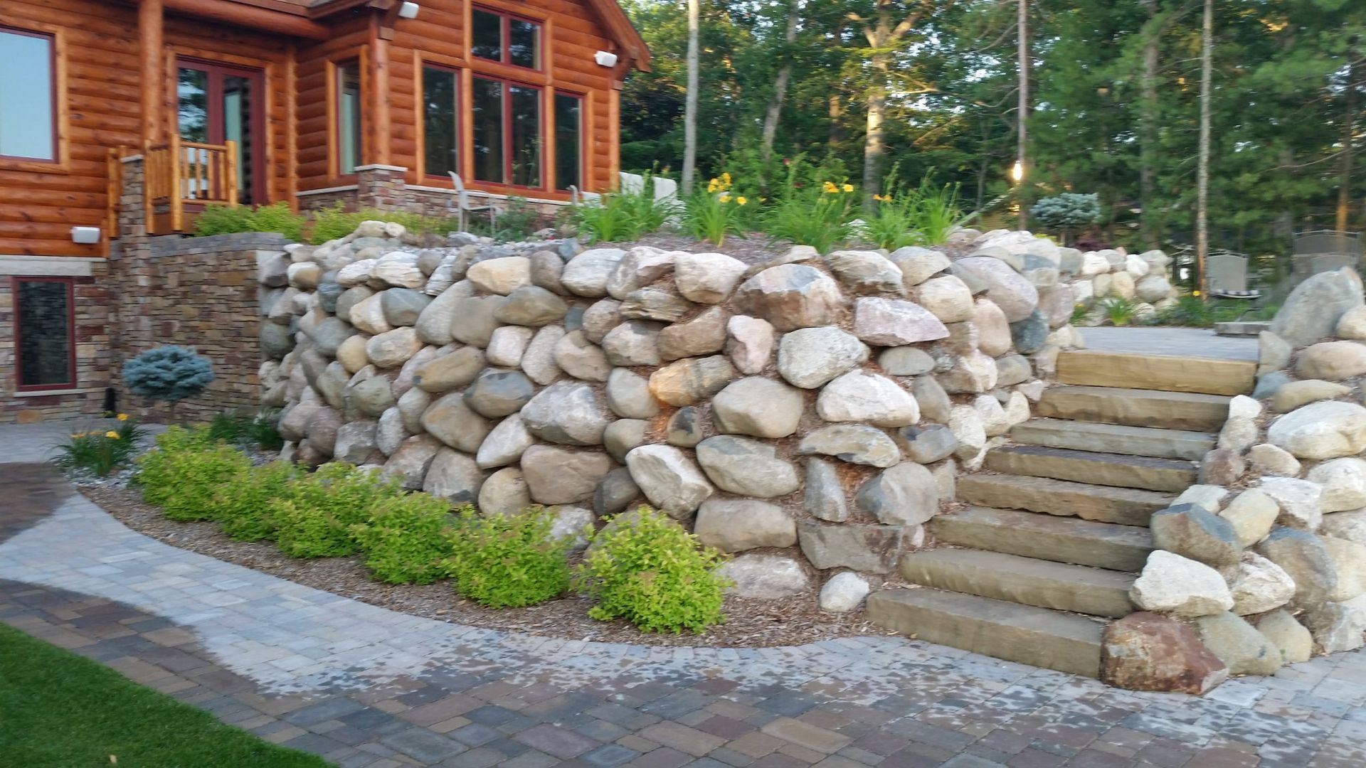 A garden path made of rectangular concrete pavers separated by rows of white decorative stones, with small green plants. Stone retaining wall with steps beside a log cabin and landscaped garden path